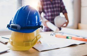 Blue and yellow hard hats on a desk beside construction plans, with a person in a plaid shirt holding a ball and a laptop in the background.